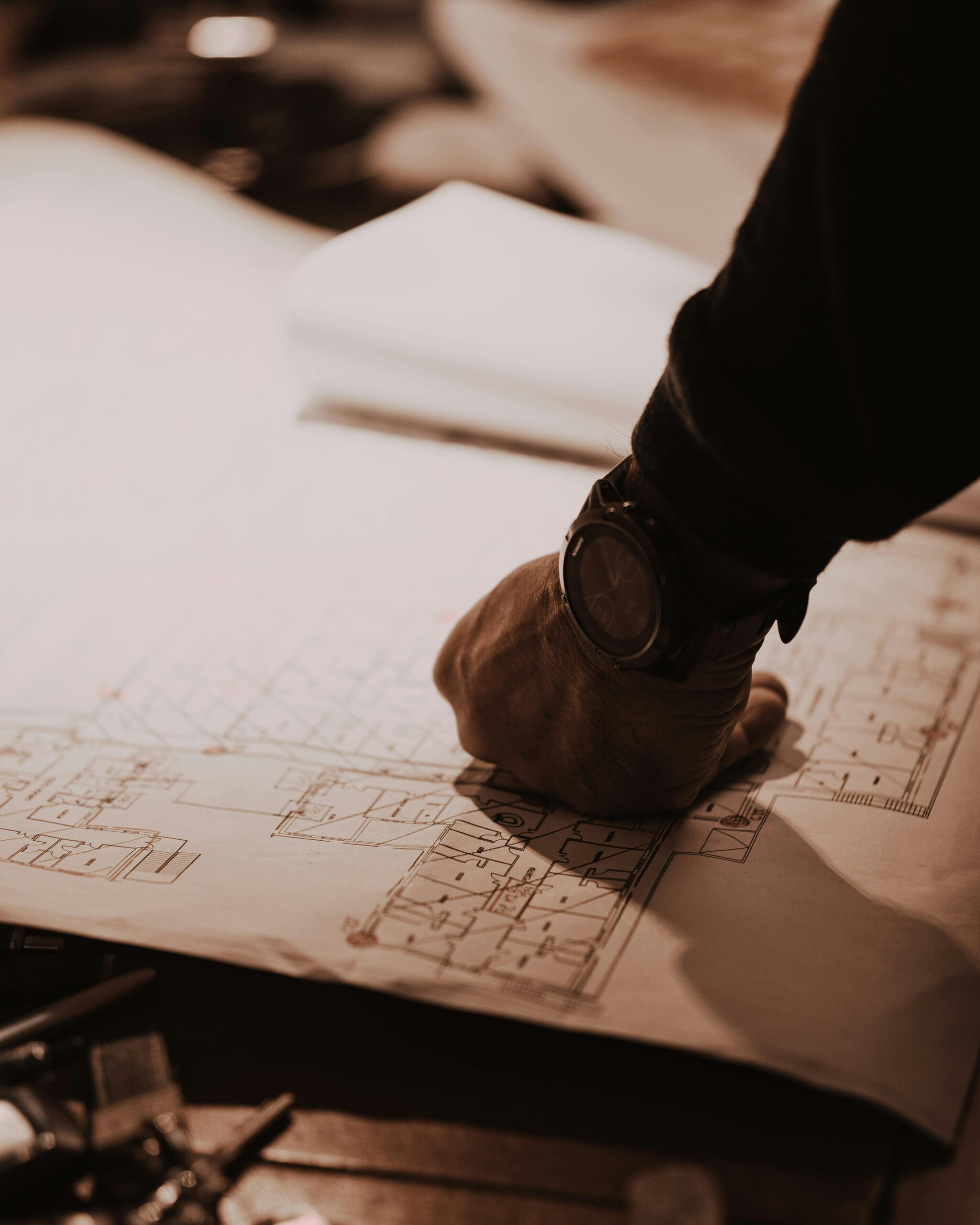 Close-up of architect's hand reviewing blueprints in a dimly lit workspace, focusing on detailed floorplans.